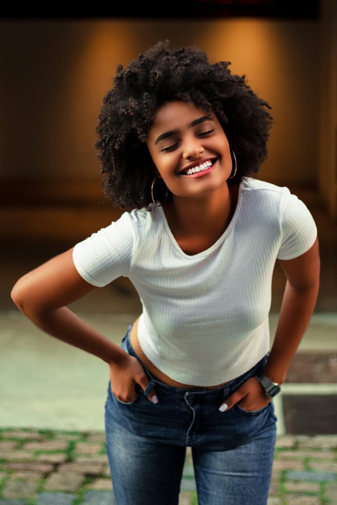 African American woman with afro hair smiling in casual white shirt and jeans, standing outdoors.